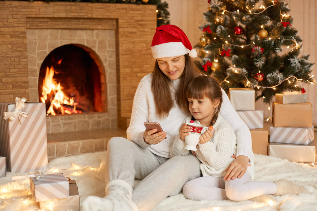 Heureuse jeune femme caucasienne avec sa petite fille qui s'amuse sur le sol, en utilisant un téléphone portable, célébrant Noël à la maison, posant près de la cheminée, enfant tenant la tasse dans les mains. - Photo, image