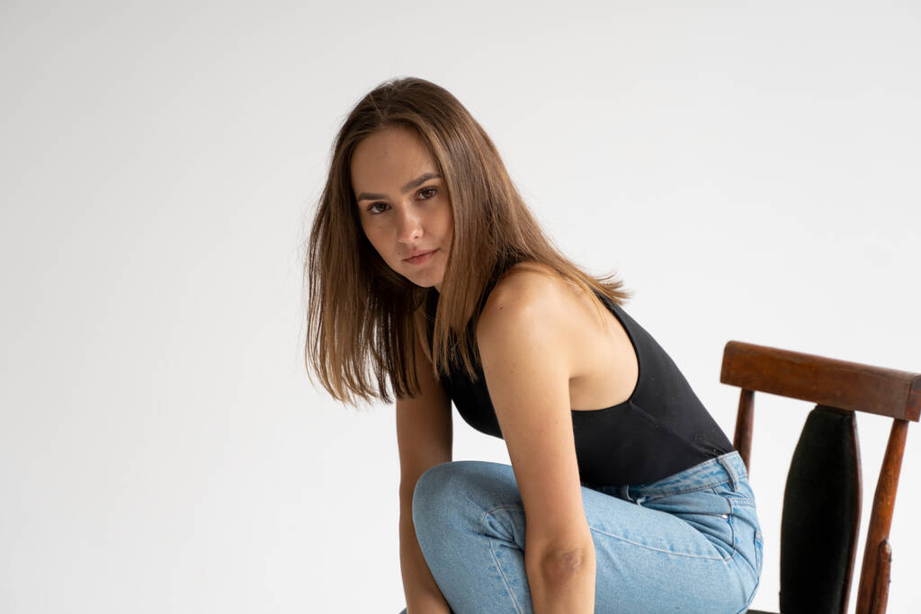 Porträt einer jungen nachdenklichen kaukasischen Frau, die in schwarzer Unterwäsche und blauer Jeans auf einem alten Stuhl im weißen Studio posiert. Modellversuche eines hübschen Mädchens in Basiskleidung auf dem Cyclorama. - Foto, Bild