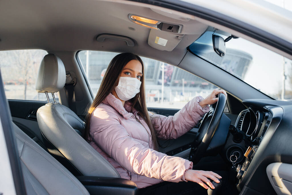 Una joven se sienta detrás del volante en el coche en la máscara durante la pandemia mundial y el coronavirus. Cuarentena - Foto, imagen