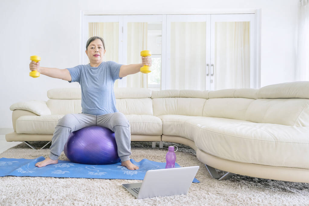 Eine ältere Frau hält zwei Kurzhanteln in der Hand und sitzt auf dem Fitnessball, während sie im Wohnzimmer trainiert. Zu Hause erschossen - Foto, Bild
