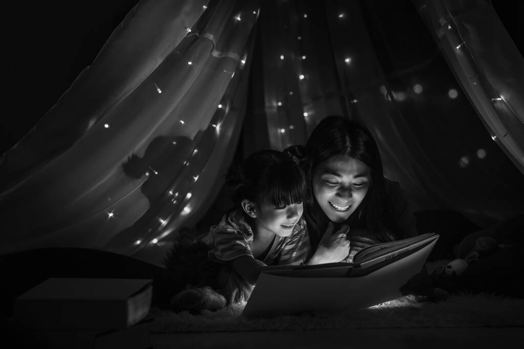 asiatische Mutter und Tochter beim gemeinsamen Lesen von Märchen für Tochter im Kinderzelt, bevor sie zu Bett gehen. Konzept der Mutter-Tochter-Beziehung in Schwarz-Weiß - Foto, Bild