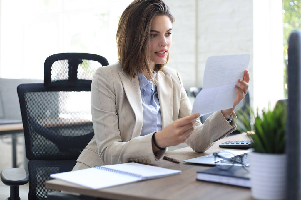 Atrakcyjny wesoły biznes kobieta sprawdzanie dokumentów papierowych w biurze, pracy na laptopie - Zdjęcie, obraz