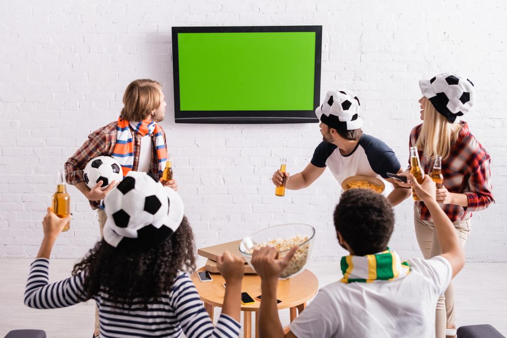 πολυπολιτισμικοί φίλοι σε ποδοσφαιρικούς οπαδούς καπέλα βλέποντας πρωτάθλημα στην τηλεόραση - Φωτογραφία, εικόνα