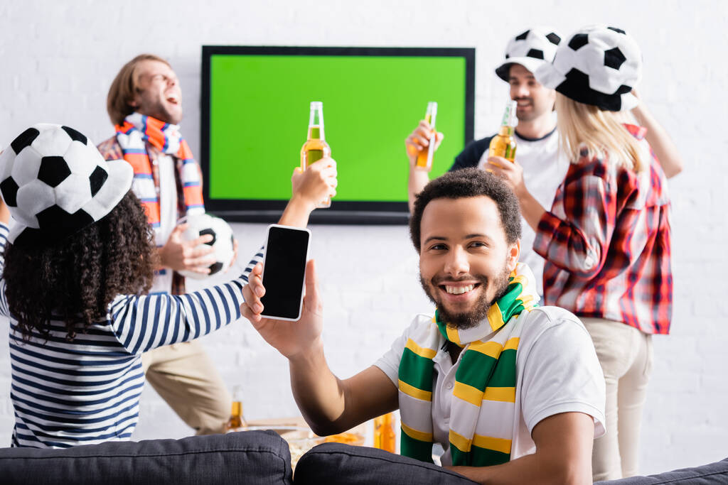veselý africký Američan ukazuje smartphone s prázdnou obrazovkou v blízkosti multikulturní přátelé ve fotbalových fanoušků klobouky na rozmazaném pozadí - Fotografie, Obrázek