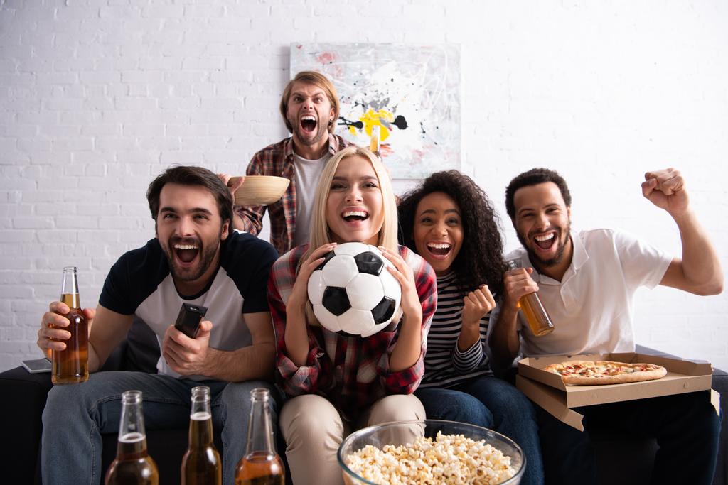 wesoła kobieta trzyma piłkę nożną podczas oglądania zawodów piłkarskich z podekscytowanymi wielonarodowymi przyjaciółmi - Zdjęcie, obraz