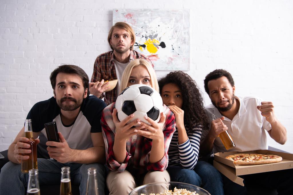 tendue femme obscurcissant visage avec ballon de football tout en regardant le championnat de football avec des amis multiculturels inquiets - Photo, image