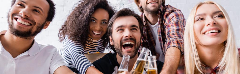 amigos multiculturales emocionados y riéndose teniendo fiesta en casa y tintineo botellas de cerveza, pancarta - Foto, Imagen
