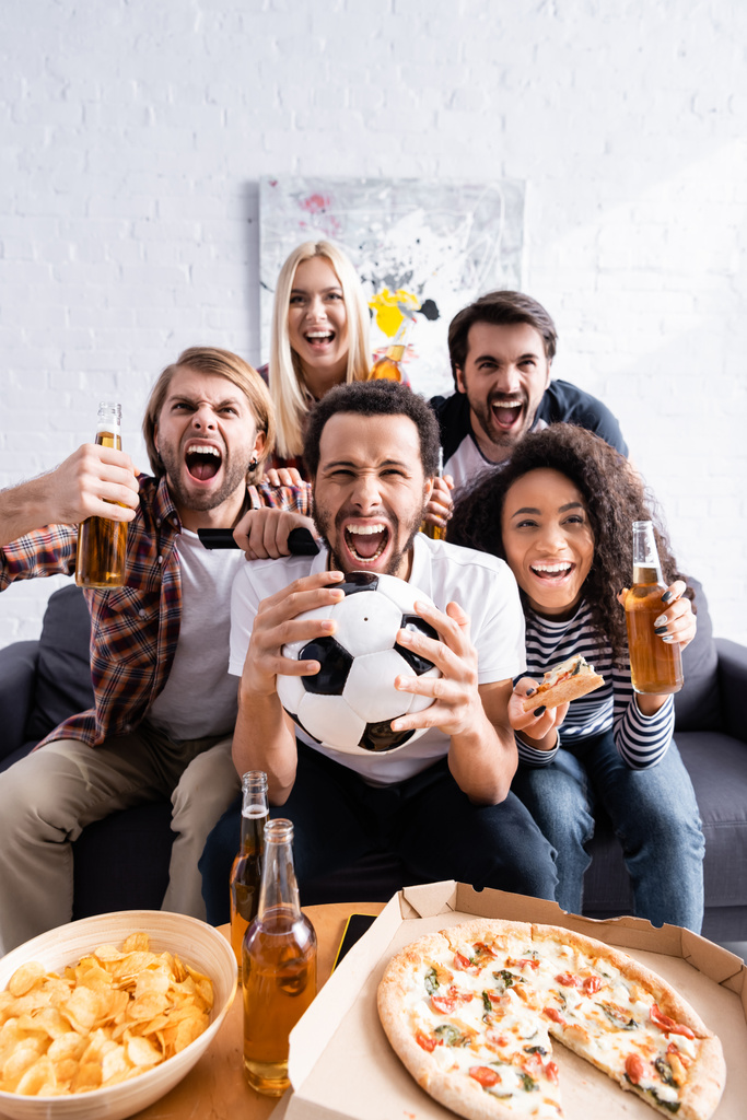 excited multiethnic football fans screaming while watching championship near beer, pizza and chips  - Photo, Image