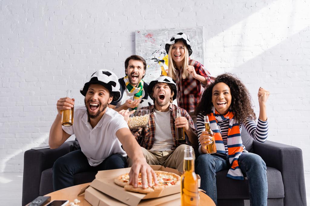 hombre emocionado tomando pizza mientras ve el campeonato de fútbol con amigos multiculturales en primer plano borrosa - Foto, imagen
