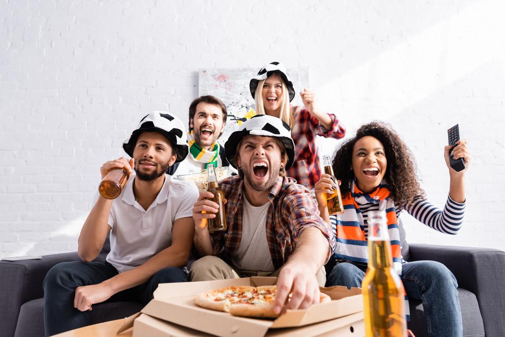 homem animado gritando, segurando cerveja e tomando pizza enquanto assiste campeonato com amigos multiculturais em primeiro plano - Foto, Imagem
