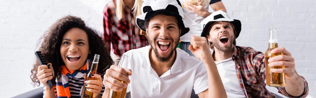 excited man screaming and showing win gesture while watching football competition with multiethnic friends on blurred background, banner - Photo, Image