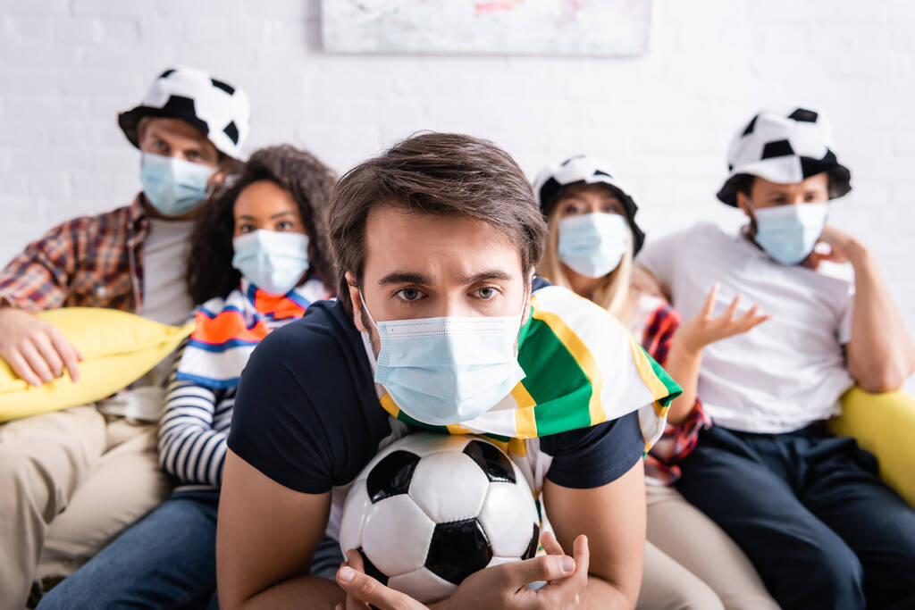 εστιασμένος άνθρωπος στην ιατρική μάσκα κρατώντας μπάλα ποδοσφαίρου, ενώ βλέποντας ποδόσφαιρο ανταγωνισμού με πολυεθνικούς φίλους σε θολή φόντο - Φωτογραφία, εικόνα