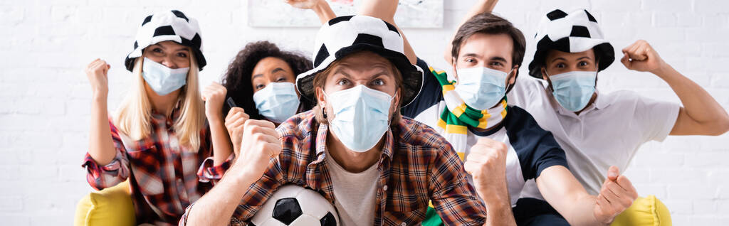 ενθουσιασμένοι πολυπολιτισμικοί φίλοι σε ποδοσφαιρικούς οπαδούς καπέλα και ιατρικές μάσκες που δείχνουν κερδίσει χειρονομία, ενώ βλέποντας πρωτάθλημα, πανό - Φωτογραφία, εικόνα