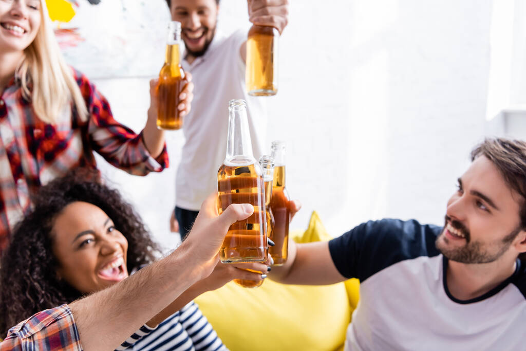 podekscytowani wielonarodowymi przyjaciółmi brzęczącymi butelkami piwa podczas imprezy, zamazane tło - Zdjęcie, obraz