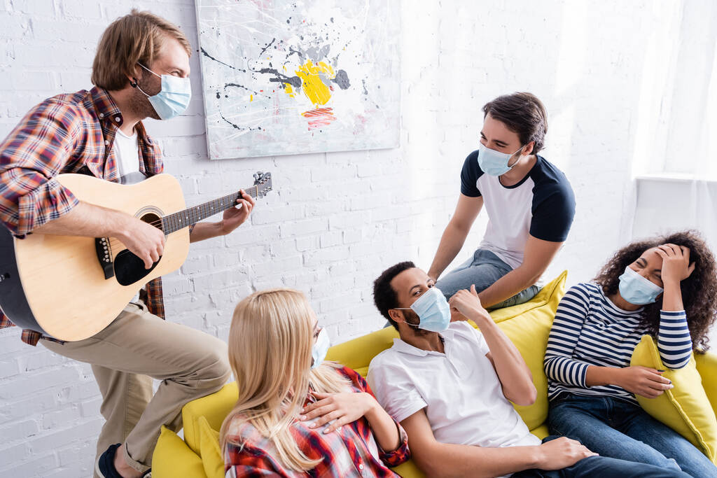 junger Mann spielt multiethnischen Freunden in medizinischen Masken Akustikgitarre während einer Party - Foto, Bild
