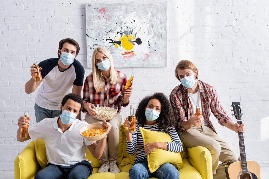 multiethnic friends in medical masks looking at camera while holding beer and snacks during party - Photo, Image