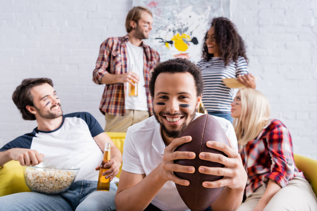 sorridente uomo africano americano che tiene palla da rugby vicino allegri amici multiculturali con facce dipinte su sfondo sfocato - Foto, immagini