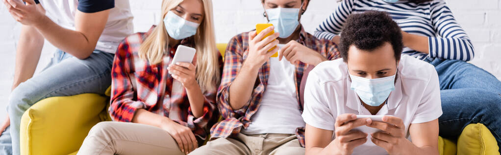 giovani amici multietnici in maschere mediche che chattano sui telefoni cellulari seduti sul divano durante la festa, sfondo sfocato, banner - Foto, immagini