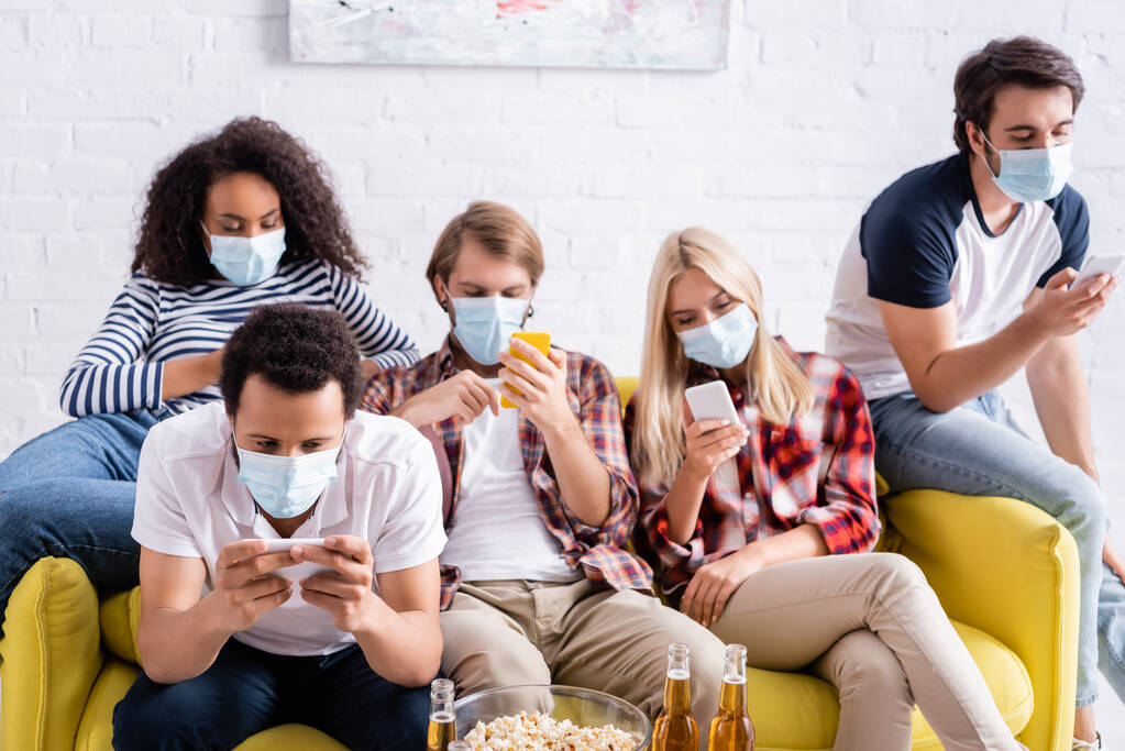 junge multiethnische Freunde in medizinischen Masken, die auf Smartphones Nachrichten senden, während sie auf dem Sofa sitzen - Foto, Bild