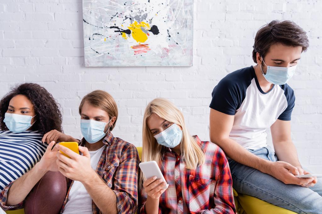 multiethnische Freunde in medizinischen Masken auf Mobiltelefonen, während sie auf dem Sofa sitzen - Foto, Bild