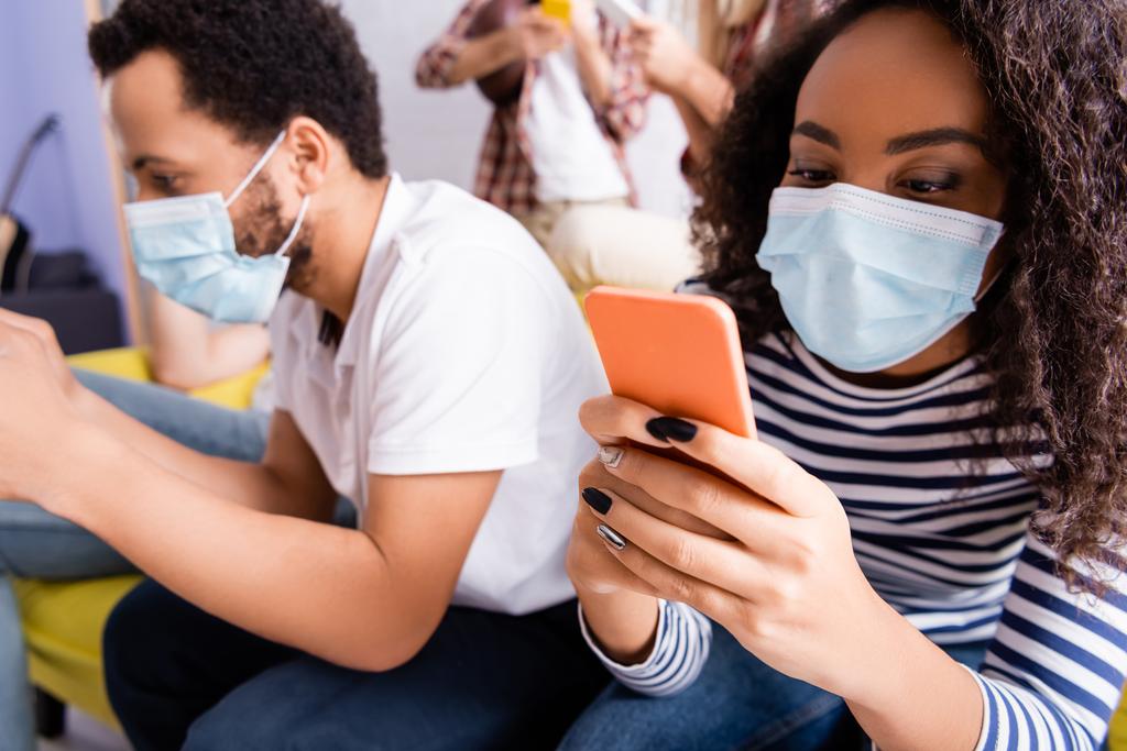 mulher americana africana em mensagens máscara médica no telefone móvel perto de amigos multiculturais em fundo turvo - Foto, Imagem