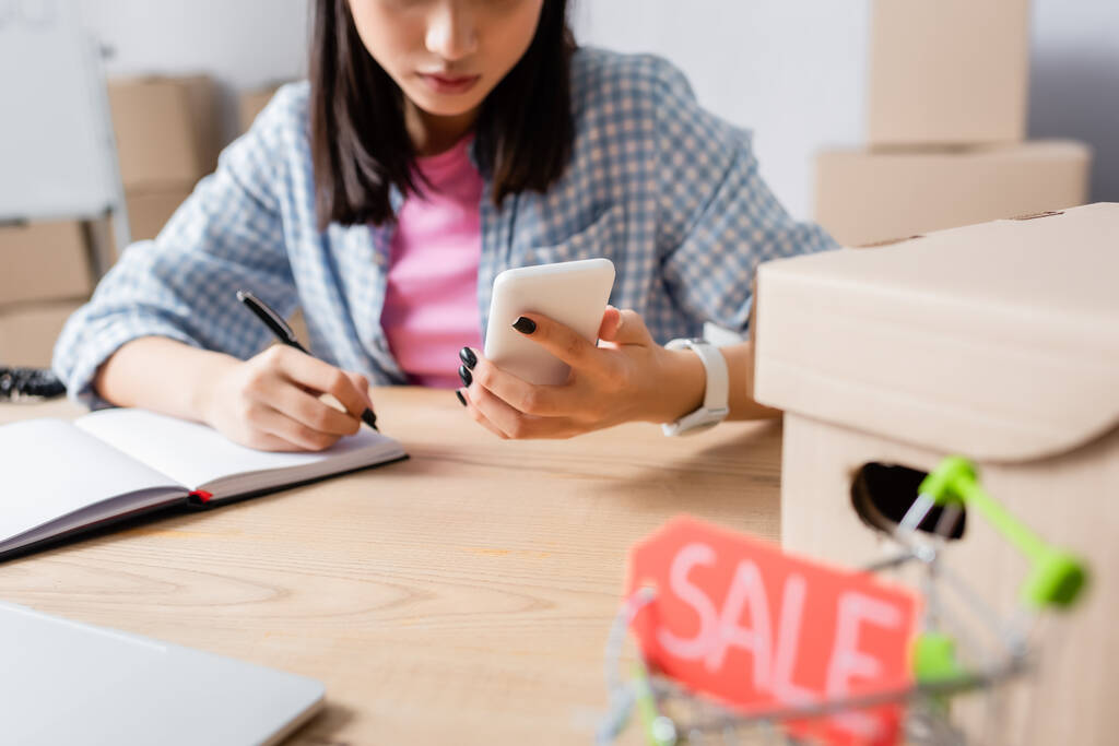 Vista recortada de voluntária feminina com escrita de smartphone em notebook na mesa com carrinho de compras borrado em primeiro plano - Foto, Imagem