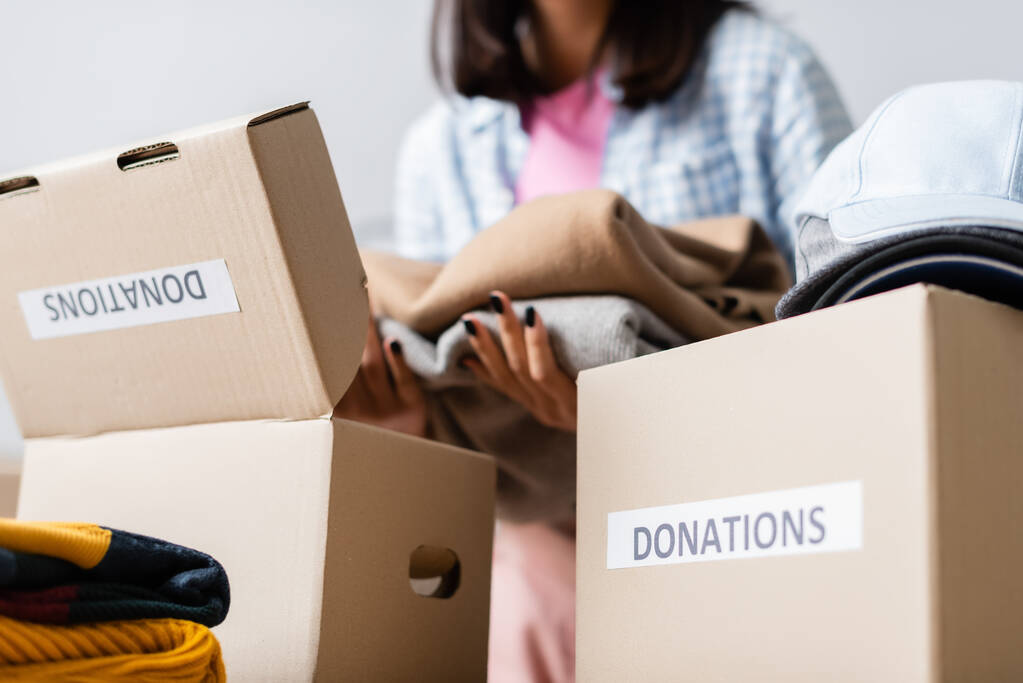 Kartonnen pakjes met donaties belettering in de buurt van jonge vrijwilliger met kleding op wazige achtergrond  - Foto, afbeelding