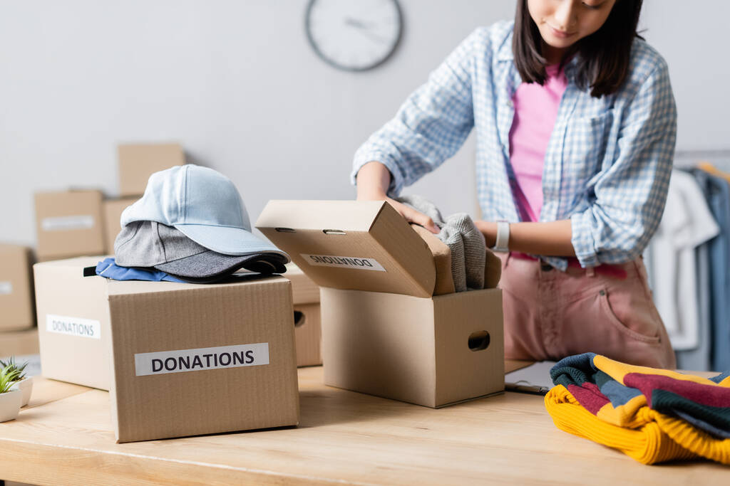 Gestroopt zicht op Aziatische vrouw met kleding in de buurt van dozen met donaties belettering en caps in het liefdadigheidscentrum  - Foto, afbeelding