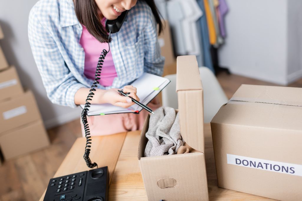 Oříznutý pohled na ženu mluvící po telefonu a ukazující na oblečení v krabici s dary v charitativním centru  - Fotografie, Obrázek