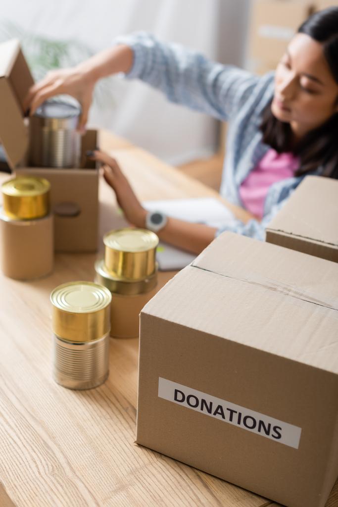 Boîte avec des dons lettrage près de la nourriture en conserve et bénévole asiatique sur fond flou  - Photo, image