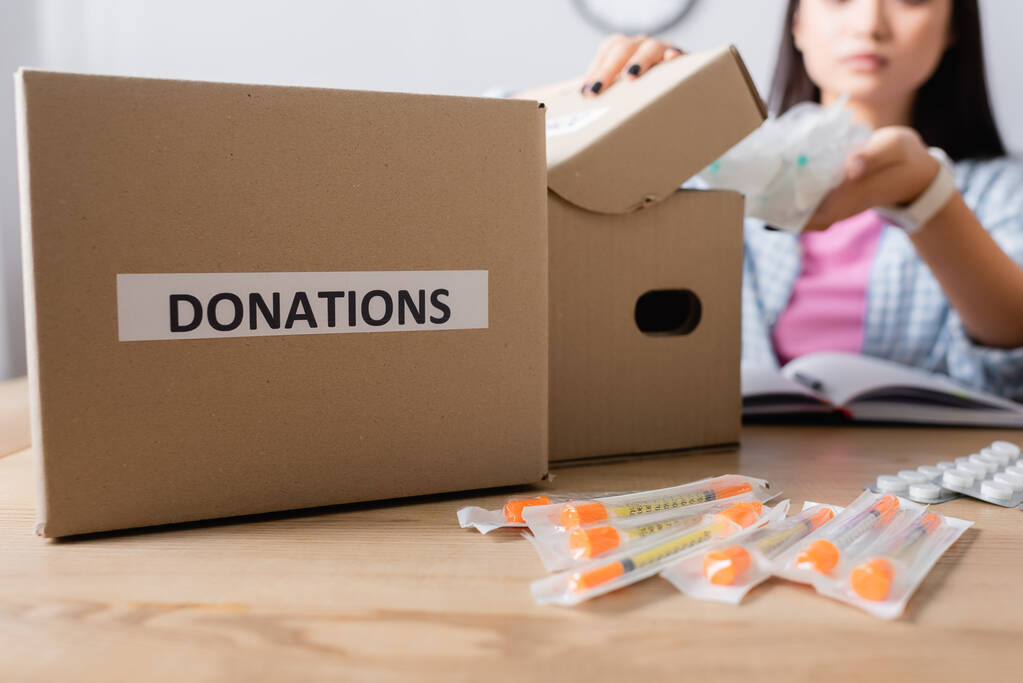 Vista recortada de la caja con letras de donaciones cerca de jeringas, pastillas y voluntarios sobre fondo borroso  - Foto, Imagen