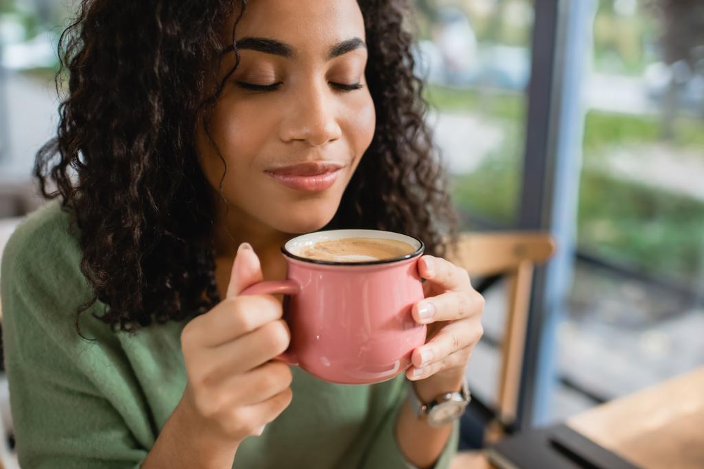 アフリカ系アメリカ人の女性がマグカップでラテコーヒーを嗅ぎ  - 写真・画像