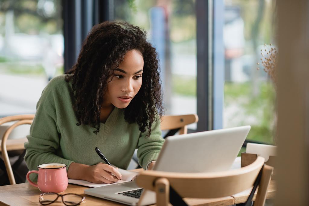 donna afro-americana che scrive nel taccuino mentre studia online nel caffè - Foto, immagini