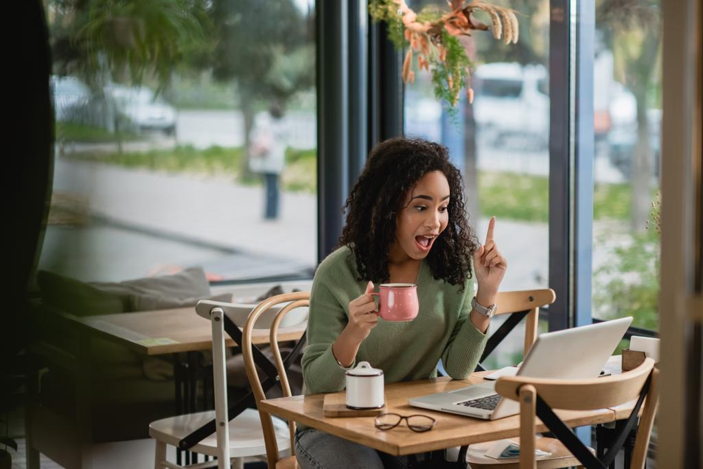 mujer afroamericana asombrada señalando con el dedo mientras que el dedo mientras que tiene idea cerca del ordenador portátil en la cafetería  - Foto, Imagen