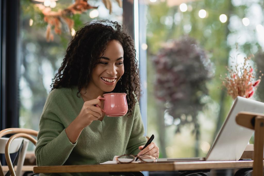 felice donna afroamericana che tiene la penna e la tazza di caffè mentre guarda il computer portatile  - Foto, immagini