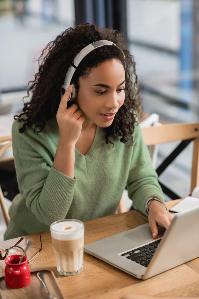 mujer afroamericana escuchando podcast cerca del ordenador portátil en la cafetería  - Foto, imagen
