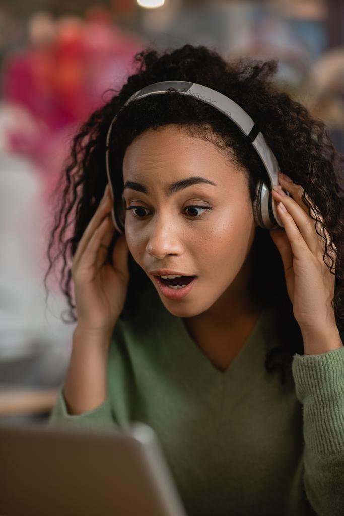 mujer afroamericana sorprendida escuchando podcast cerca de la computadora portátil en la cafetería  - Foto, imagen