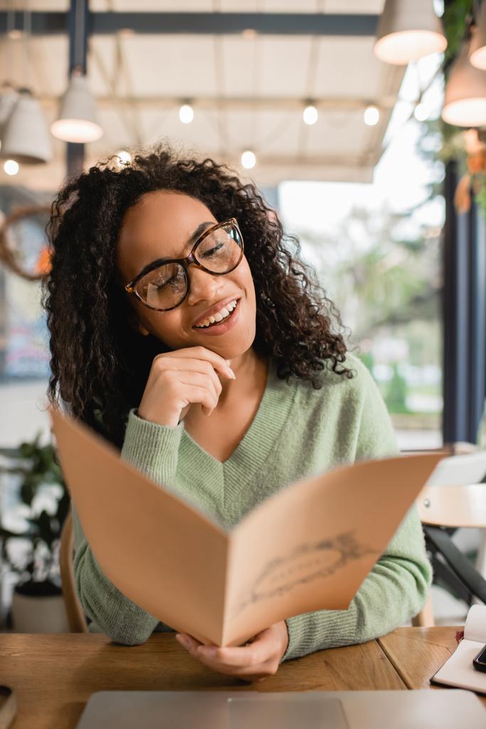 happy curly african american woman in glasses holding menu on blurred foreground  - Photo, Image