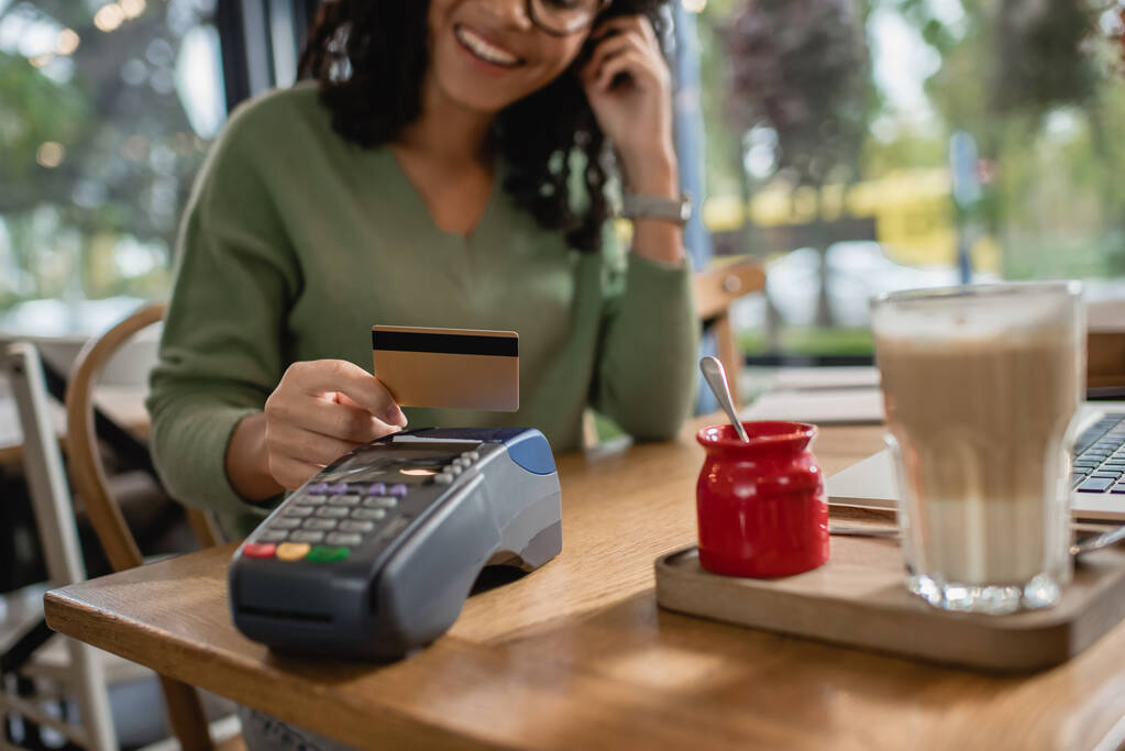 vista recortada de la mujer afroamericana feliz sosteniendo tarjeta de crédito por encima de la terminal de pago en la cafetería - Foto, Imagen