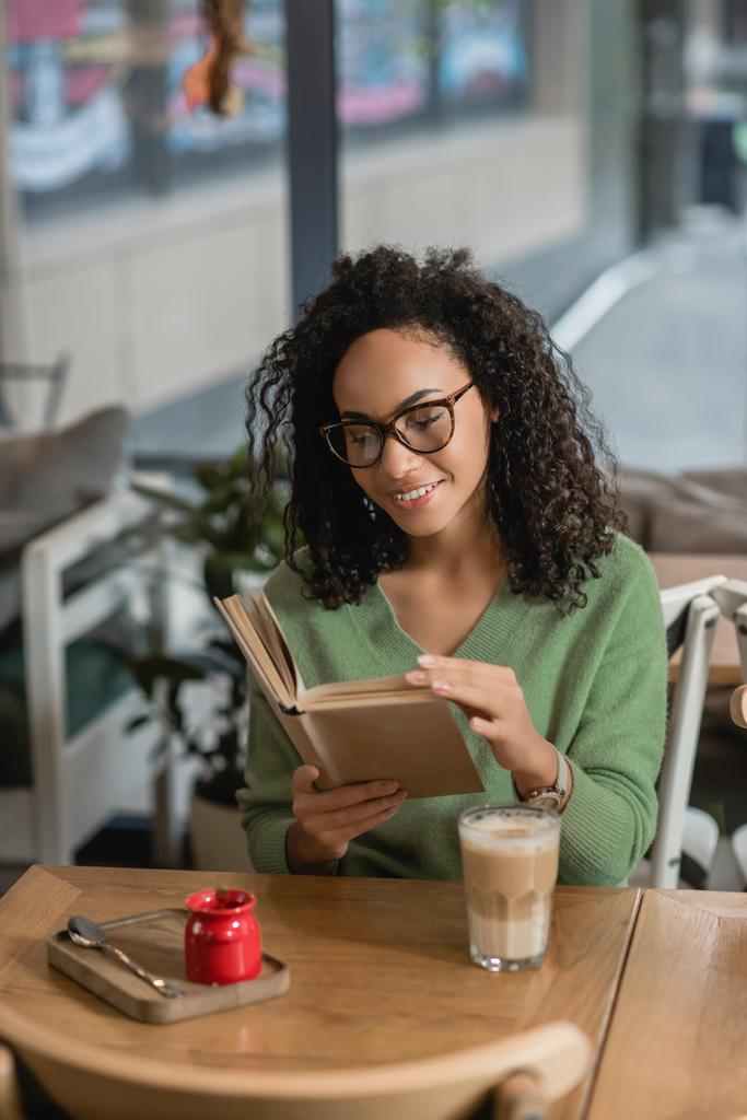 happy african american woman in eyeglasses reading book near glass with latte - Photo, Image