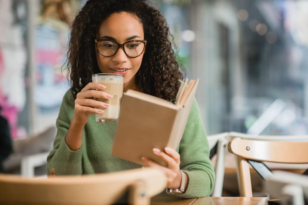 alegre afroamericana mujer leyendo libro y sosteniendo vidrio con café con leche en la cafetería - Foto, imagen