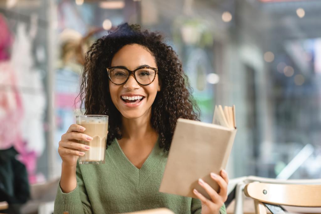 sonriente mujer afroamericana sosteniendo libro y vidrio con café con leche en la cafetería - Foto, Imagen