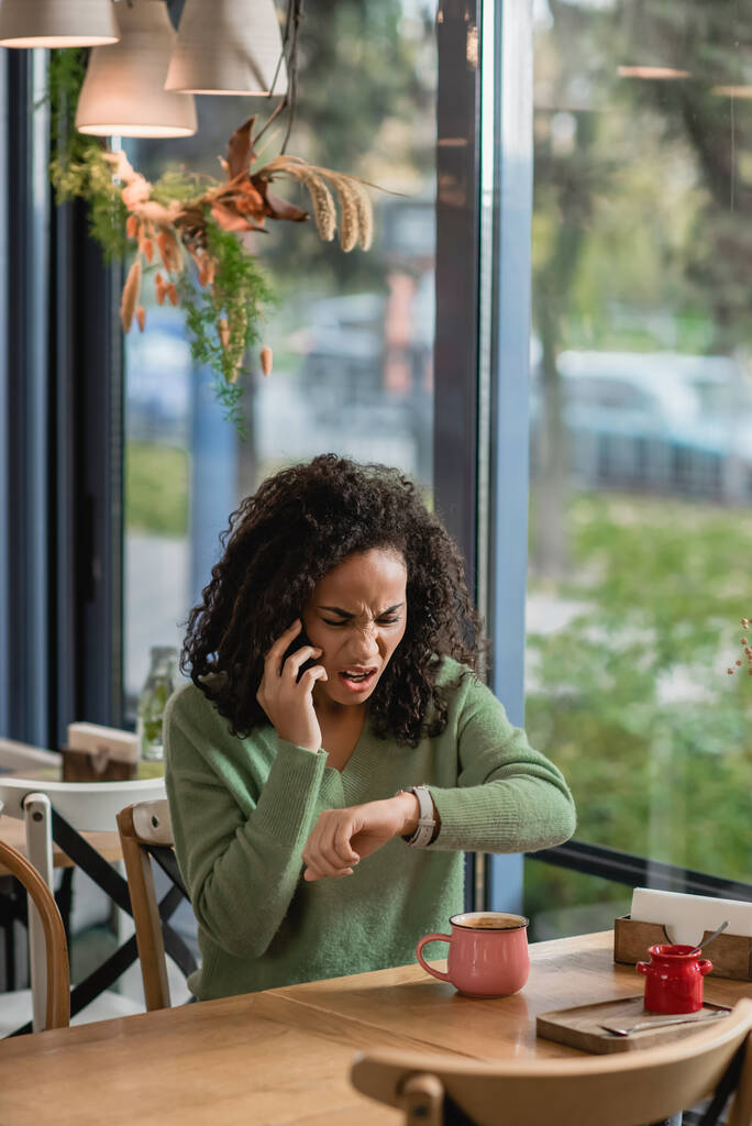 boos Afrikaans amerikaanse vrouw praten op smartphone, terwijl het controleren van de horloge op de pols en wachten in cafe - Foto, afbeelding