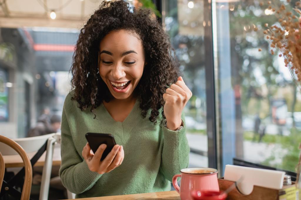 excitée femme afro-américaine avec poing serré regardant smartphone dans le café - Photo, image