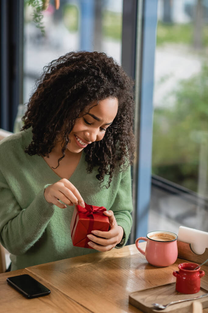 mujer afroamericana feliz tirando de la cinta en la caja de regalo de Navidad roja en la cafetería - Foto, Imagen