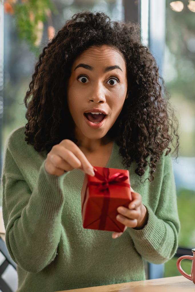 σοκαρισμένη Αφροαμερικανή τραβώντας κορδέλα σε κόκκινο κουτί δώρου Χριστουγέννων στο καφέ - Φωτογραφία, εικόνα