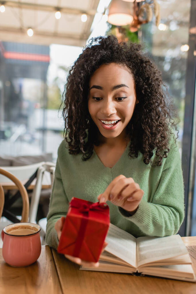 szczęśliwy Afryki amerykanka ciągnąc wstążkę na czerwony świąteczny prezent pudełko w kawiarni - Zdjęcie, obraz