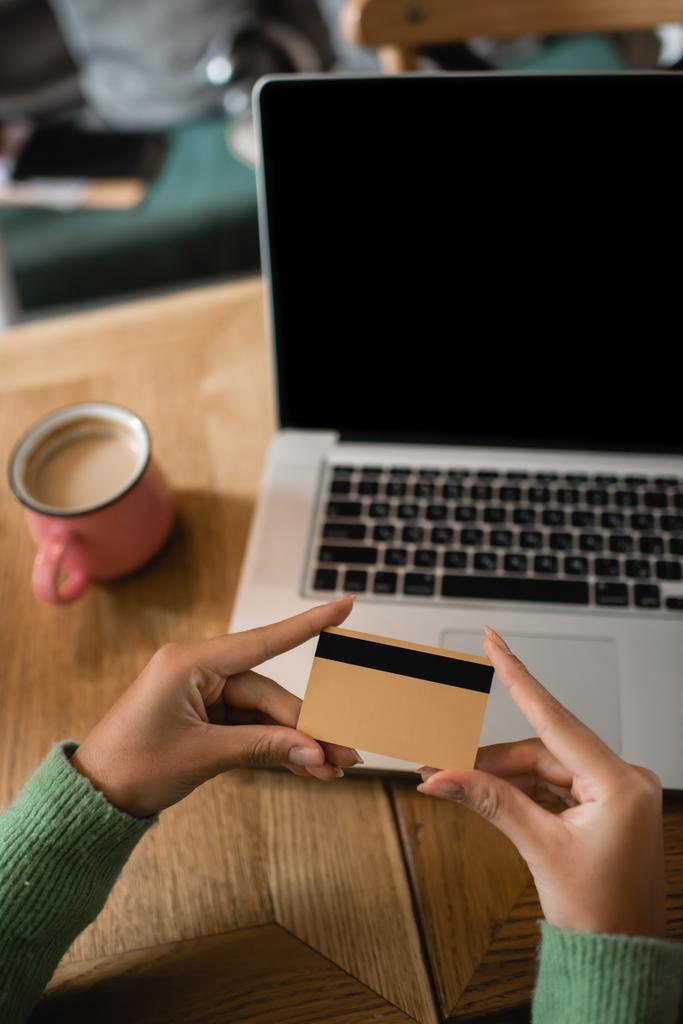 vista recortada de la mujer afroamericana que sostiene la tarjeta de crédito cerca del ordenador portátil con la pantalla en blanco  - Foto, imagen
