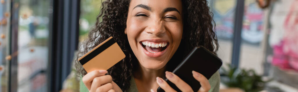 cheerful african american woman laughing while holding smartphone and credit card, banner - Photo, Image