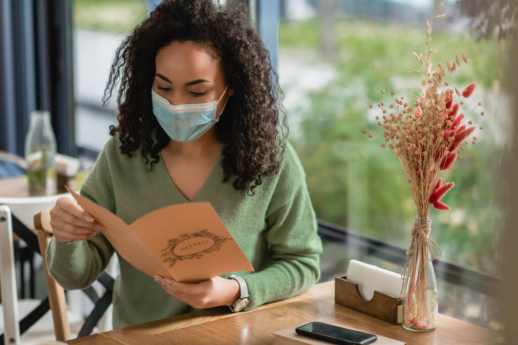 donna afroamericana in maschera medica che tiene il menu nel caffè - Foto, immagini